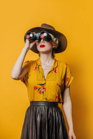 Foto de Elegante mujer de pelo rubio en sombrero y camisa con prismáticos sobre fondo amarillo - Imagen libre de derechos