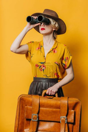 Foto de Elegante mujer de pelo rubio en sombrero y camisa con prismáticos y maleta sobre fondo amarillo - Imagen libre de derechos