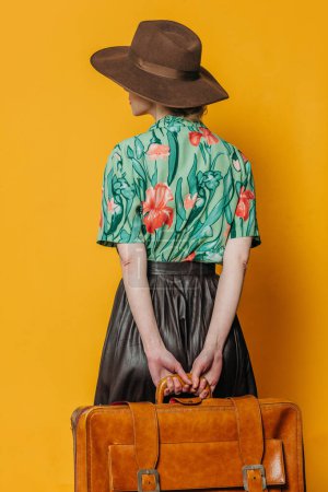 Téléchargez les photos : Vue du dos sur une élégante femme blonde coiffée d'un chapeau et d'une chemise avec une valise sur fond jaune - en image libre de droit