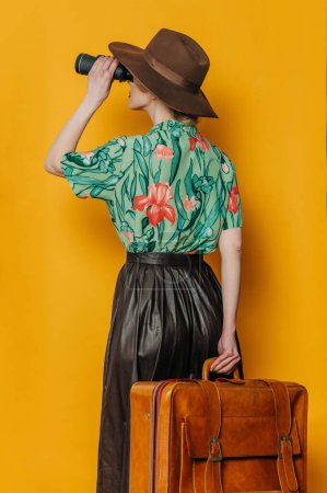Foto de Vista lateral posterior sobre elegante mujer rubia de pelo en sombrero y camisa con prismáticos y maleta sobre fondo amarillo - Imagen libre de derechos
