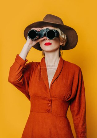 Foto de Elegante mujer de pelo rubio en sombrero y vestido marrón con binoculares sobre fondo amarillo - Imagen libre de derechos