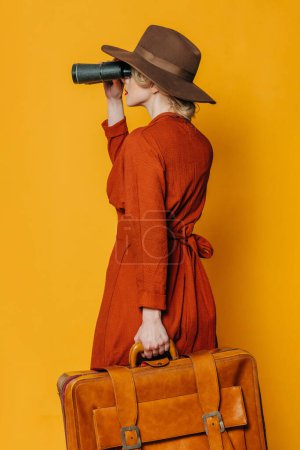 Foto de Vista lateral trasera de la mujer en sombrero y vestido marrón con maleta y binoculares sobre fondo amarillo - Imagen libre de derechos