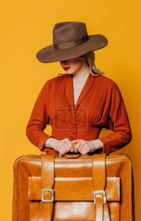 Foto de Mujer de pelo rubio en sombrero vintage y vestido marrón con maleta sobre fondo amarillo - Imagen libre de derechos