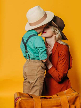 Foto de Elegante madre e hijo en ropa vintage con maleta sobre fondo amarillo - Imagen libre de derechos