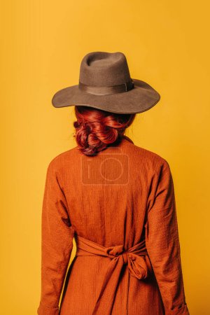 Téléchargez les photos : Vue de face arrière sur femme rousse élégante en chapeau et robe brune sur fond jaune - en image libre de droit