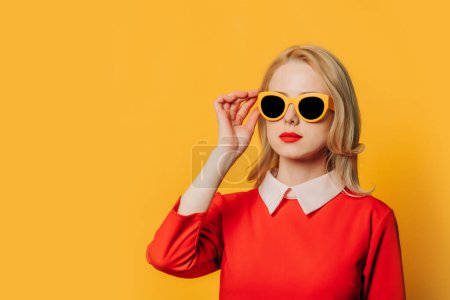 Foto de Elegante mujer de pelo rubio serio en vestido rojo y gafas de sol sobre fondo amarillo - Imagen libre de derechos