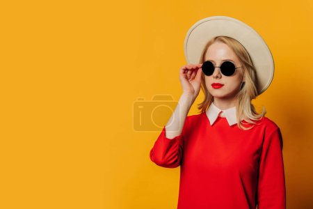 Foto de Elegante mujer de pelo rubio en vestido rojo y gafas de sol sobre fondo amarillo - Imagen libre de derechos