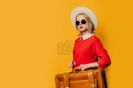 Foto de Elegante mujer de pelo rubio en vestido rojo con maleta sobre fondo amarillo - Imagen libre de derechos