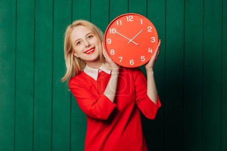 Téléchargez les photos : Femme aux cheveux blonds souriants en robe rouge avec horloge sur fond de bois vert - en image libre de droit