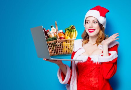 Photo for Portrait of Young Santa Clous girl in red clothes with laptop computer and basket full of products on blue background - Royalty Free Image