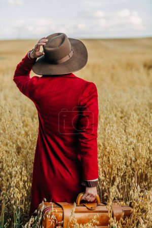 Photo for Stylish woman in vintage hat red coat with suitcase on oat field in countryside - Royalty Free Image