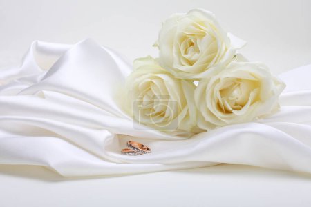Téléchargez les photos : Nacre wave fabric silk with white rose flower bouquet and wedding ring. Abstract texture horizontal copy space background. - en image libre de droit