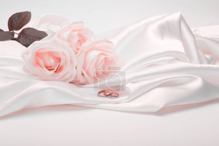 Téléchargez les photos : Nacre wave fabric silk with beige pink rose flower bouquet and wedding ring. Abstract texture horizontal copy space background. - en image libre de droit