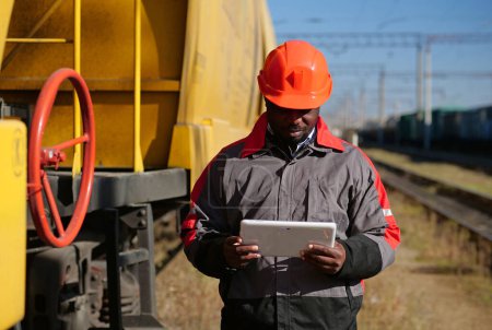 Photo for Inspector of wagons at freight train station makes notes in Tablet PC. African american railway man with tablet computer at freight train terminal - Royalty Free Image
