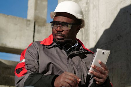 Foto de Obrero afroamericano se sienta en las escaleras en el sitio de construcción y sostiene en las manos smartphone - Imagen libre de derechos