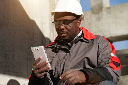 Photo for African american workman sits on the stairs at construction site and looks in smartphon - Royalty Free Image