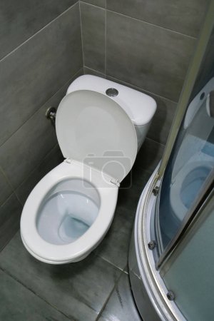 Toilet room interior with white toilet bowl, shower cabin and grey wall tiles