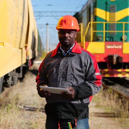 Photo for Railway employee holds in hands tablet pc and look at camera. Railroad man in uniform and red hard hat with computer. African american railway man with tablet computer at freight train terminal - Royalty Free Image
