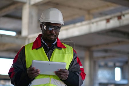 Téléchargez les photos : Travailleur afro-américain en casque dur et lunettes de protection se tient sur le chantier de construction, tient dans les mains tablette ordinateur et regarde la caméra - en image libre de droit