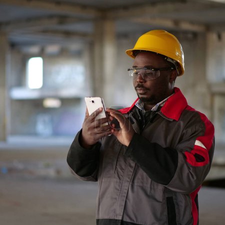 Photo for African american workman in yellow hard hat with smartphone stands at construction site. Worker holds in hands smartphone - Royalty Free Image