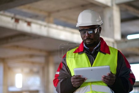 Téléchargez les photos : Travailleur afro-américain en casque dur et lunettes de protection avec les yeux fermés se tient sur le chantier de construction, tient dans les mains Tablette PC - en image libre de droit