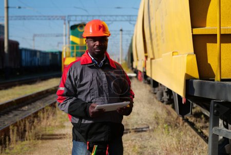 Photo for Railroad man in uniform and red hard hat with computer. African american railway man with tablet computer at freight train terminal. Railway employee holds in hands tablet pc and look at camera - Royalty Free Image