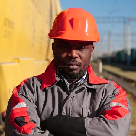 Photo for Railroad man in uniform and red hard hat stands on railroad tracks. African american railway worker patrol lineman stands at freight train terminal and looks at the camera - Royalty Free Image