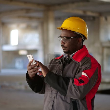 African american workman in yellow hard hat with smartphone stands at construction site. Worker holds in hands smartphone