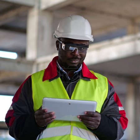 Téléchargez les photos : Travailleur afro-américain en casque dur et lunettes de protection se tient sur le chantier de construction, tient dans les mains tablette ordinateur, regarde la caméra et sourit - en image libre de droit