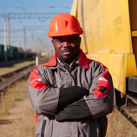 Photo for African american railway worker stands at freight train terminal and looks at camera. Railroad man in uniform and red hard hat stands on railroad tracks - Royalty Free Image