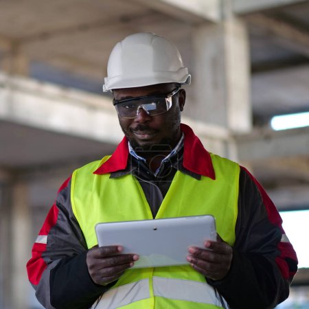 Téléchargez les photos : Travailleur afro-américain en casque dur et lunettes de protection se tient sur le chantier de construction, tient dans les mains tablette ordinateur, regarde la caméra et sourit - en image libre de droit