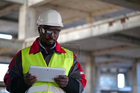 Téléchargez les photos : Travailleur afro-américain en casque dur et lunettes de protection avec les yeux fermés se tient sur le chantier de construction, tient dans les mains Tablette PC - en image libre de droit