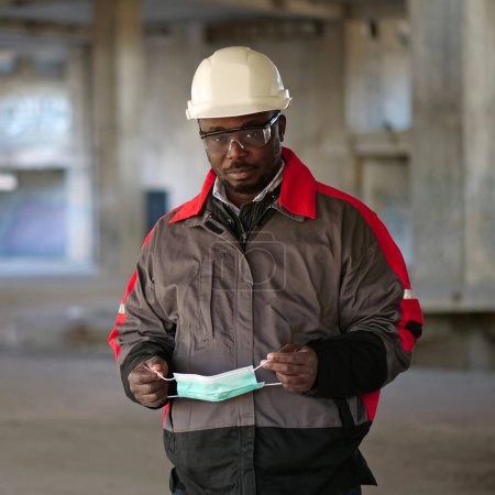 Téléchargez les photos : Constructeur afro-américain se tient sur le chantier de construction, regarde la caméra et tient dans les mains masque de protection médicale - en image libre de droit
