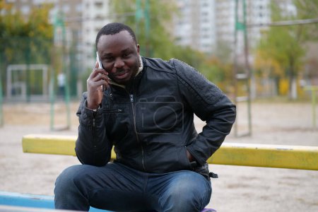 Téléchargez les photos : L'homme afro-américain est assis au terrain de sport, parle sur smartphone, regarde la caméra et sourit - en image libre de droit
