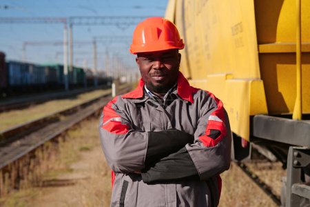 Photo for African american railway worker stands at freight train terminal and looks at camera. Railroad man in uniform and red hard hat stands on railroad tracks - Royalty Free Image