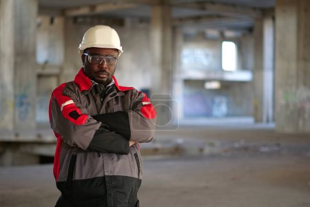 Photo for African american worker in white hard hat and protective goggles stands at construction area and looks at the camera - Royalty Free Image
