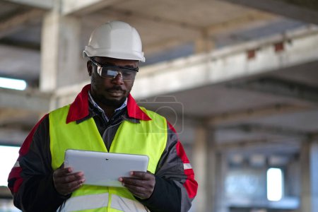 Téléchargez les photos : Travailleur afro-américain en casque dur et lunettes de protection avec les yeux fermés se tient sur le chantier de construction, tient dans les mains tablette ordinateur - en image libre de droit