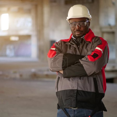 Photo for African american worker in white hard hat and protective goggles stands at construction area and looks at the camera - Royalty Free Image
