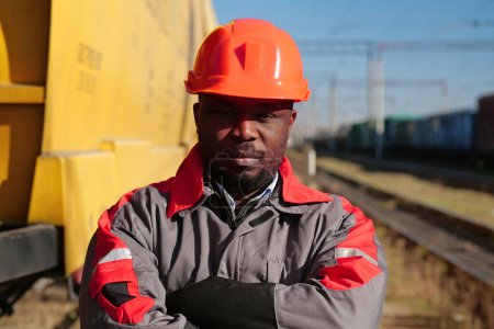 Photo for Railroad man in uniform and red hard hat stands on railroad tracks. African american railway worker patrol lineman stands at freight train terminal and looks at the camera - Royalty Free Image