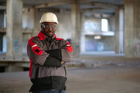 Photo for African american worker in white hard hat and protective goggles stands at construction area, looks at the camera and smiles - Royalty Free Image