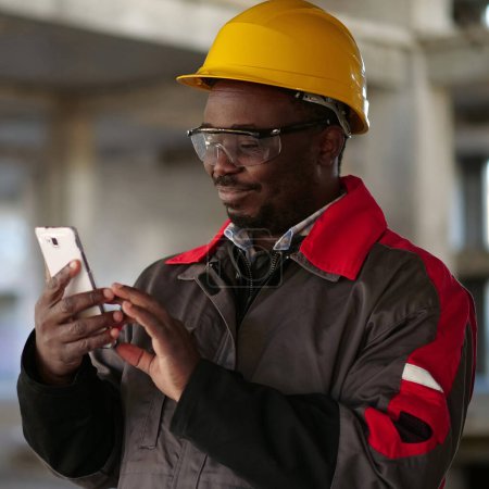 Téléchargez les photos : Un ouvrier afro-américain en casque jaune avec smartphone se tient sur le chantier. Travailleur tient dans les mains smartphone - en image libre de droit