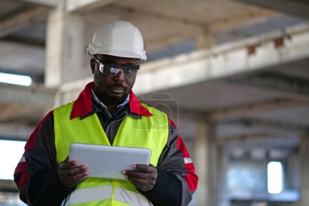 Téléchargez les photos : Travailleur afro-américain en casque dur et lunettes de protection se tient sur le chantier de construction, tient dans les mains tablette ordinateur et regarde la caméra - en image libre de droit