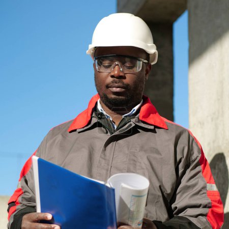 Téléchargez les photos : Un ouvrier afro-américain se tient sur le chantier et regarde les papiers de travail - en image libre de droit
