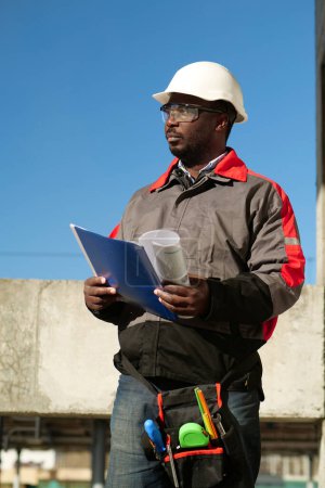 Téléchargez les photos : Un ouvrier afro-américain se tient sur le chantier avec des papiers de travail - en image libre de droit