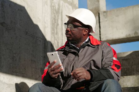 Téléchargez les photos : Travailleur afro-américain en casque dur et lunettes de protection avec smartphone se trouve sur le chantier de construction. Workman tient dans les mains smartphone blanc et regarde autour de lui - en image libre de droit
