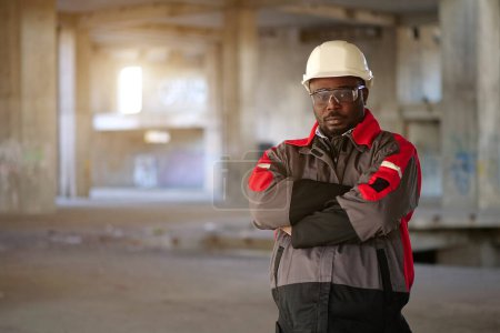 Téléchargez les photos : Travailleur afro-américain en casque blanc et lunettes de protection se tient à la zone de construction et regarde la caméra - en image libre de droit