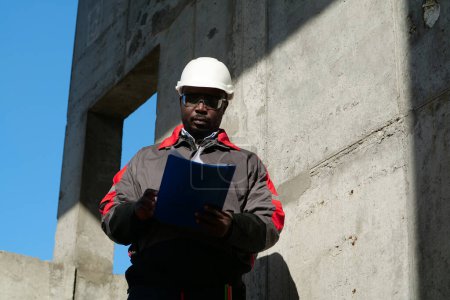 Téléchargez les photos : Un ouvrier afro-américain se tient sur le chantier avec des papiers de travail - en image libre de droit