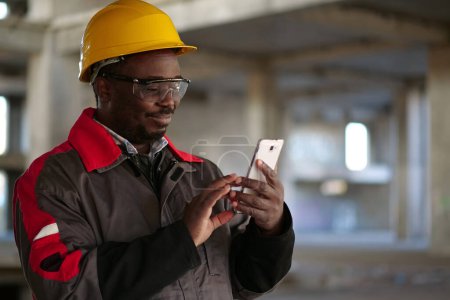 Photo for African american workman in yellow hard hat with smartphone stands at construction site. Worker holds in hands smartphone - Royalty Free Image