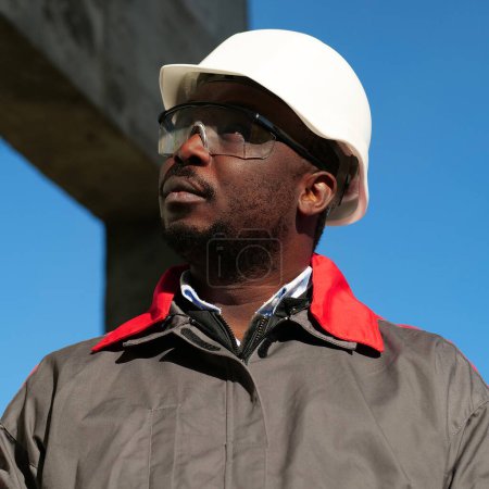 Téléchargez les photos : Travailleur afro-américain en casque et lunettes de protection se tient sur le chantier de construction - en image libre de droit