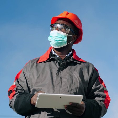 Téléchargez les photos : Travailleur afro-américain dans le masque de protection médicale se tient à la construction avec tablette informatique. Workman tient dans les mains une tablette PC blanche et regarde autour de lui - en image libre de droit